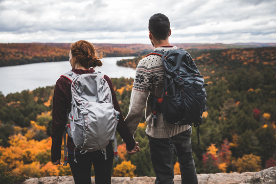 Un couple en randonnée sur une colline rocheuse admire les couleurs automnales de la forêt en contrebas.